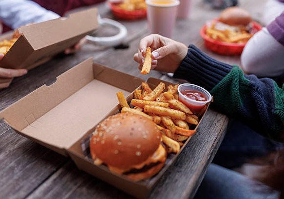 Close up of teenage girl sitting in fast food restaurant outdoors and holding french fries, there is a burger with french fries on a table